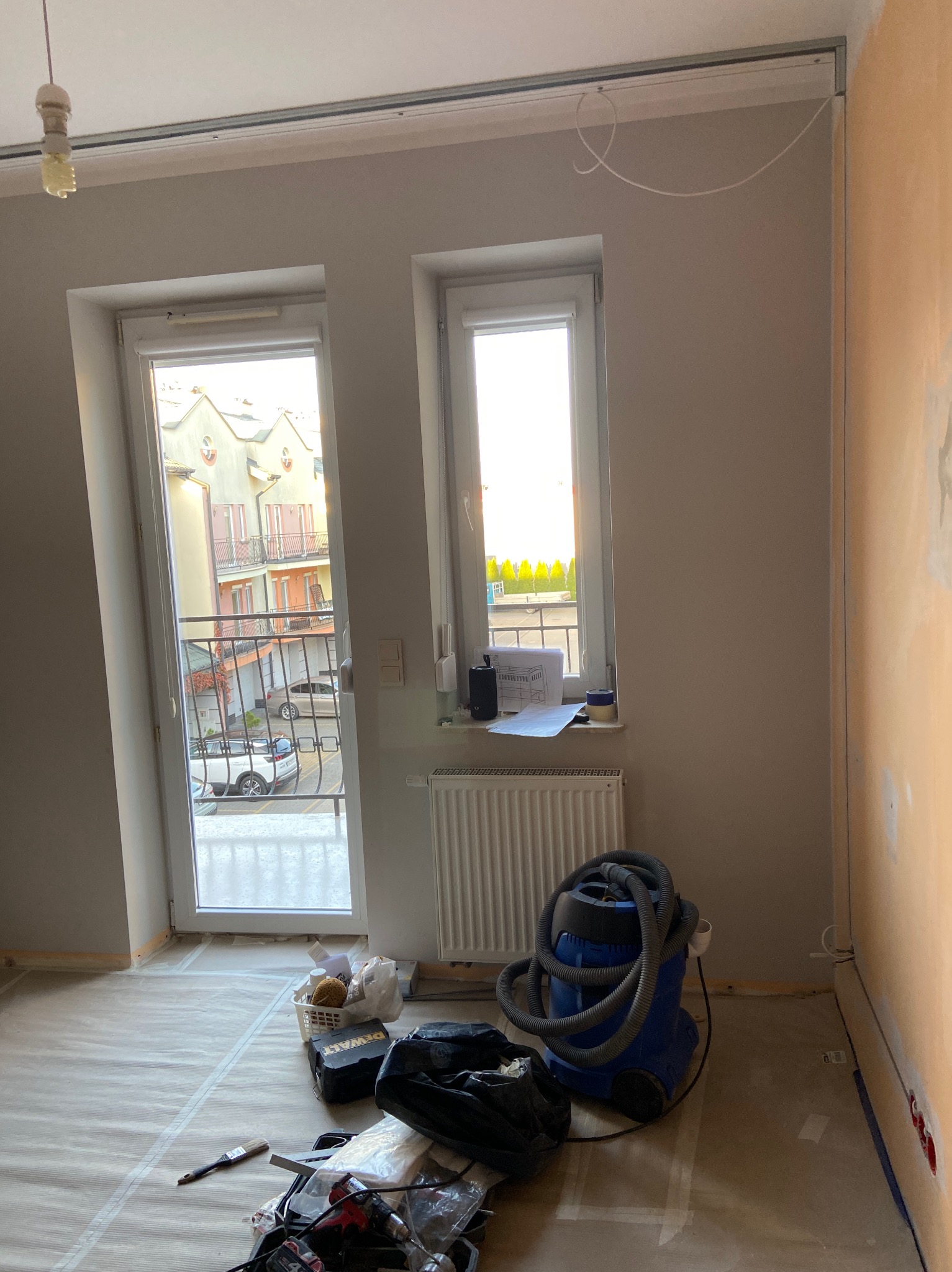 Pokój w trakcie przygotowania do remontu: zabezpieczona podłoga folią malarską, sprzęt i narzędzia budowlane, ściana częściowo przygotowana do malowania, widok na balkon i okno.