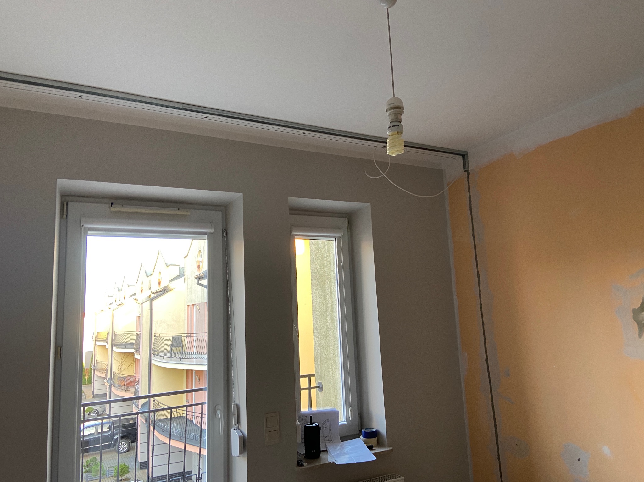 Wnętrze pokoju w trakcie przygotowań do malowania: widoczne okna z widokiem na budynek z balkonami, ściana pomalowana częściowo na żółto z widocznymi śladami po szpachlowaniu, listwa sufitowa...