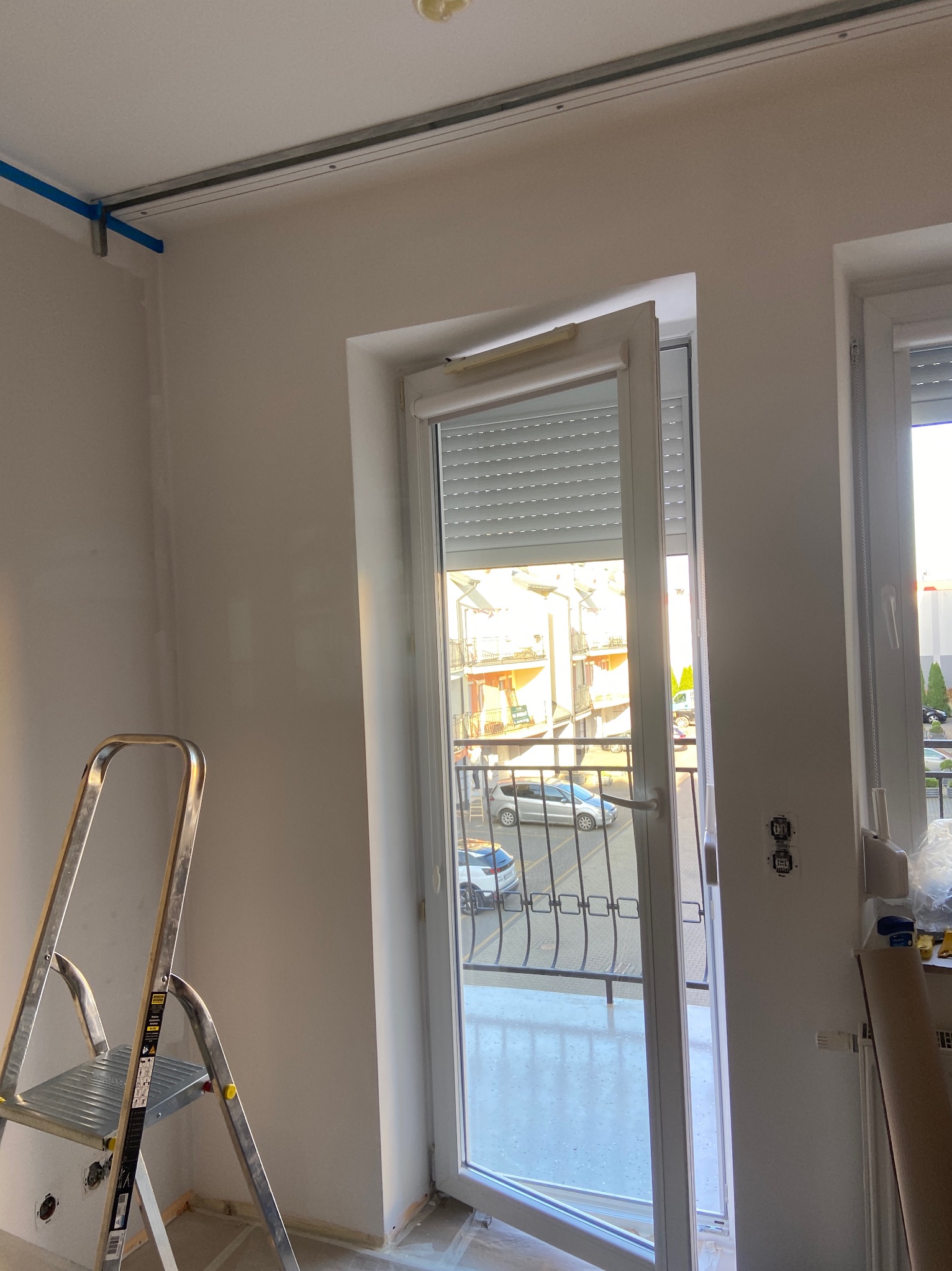 Wnętrze w trakcie odświeżania: metalowa drabina, zabezpieczone taśmą sufitowe szyny, otwarte okno balkonowe z roletami zewnętrznymi, widoczne gniazdka elektryczne.