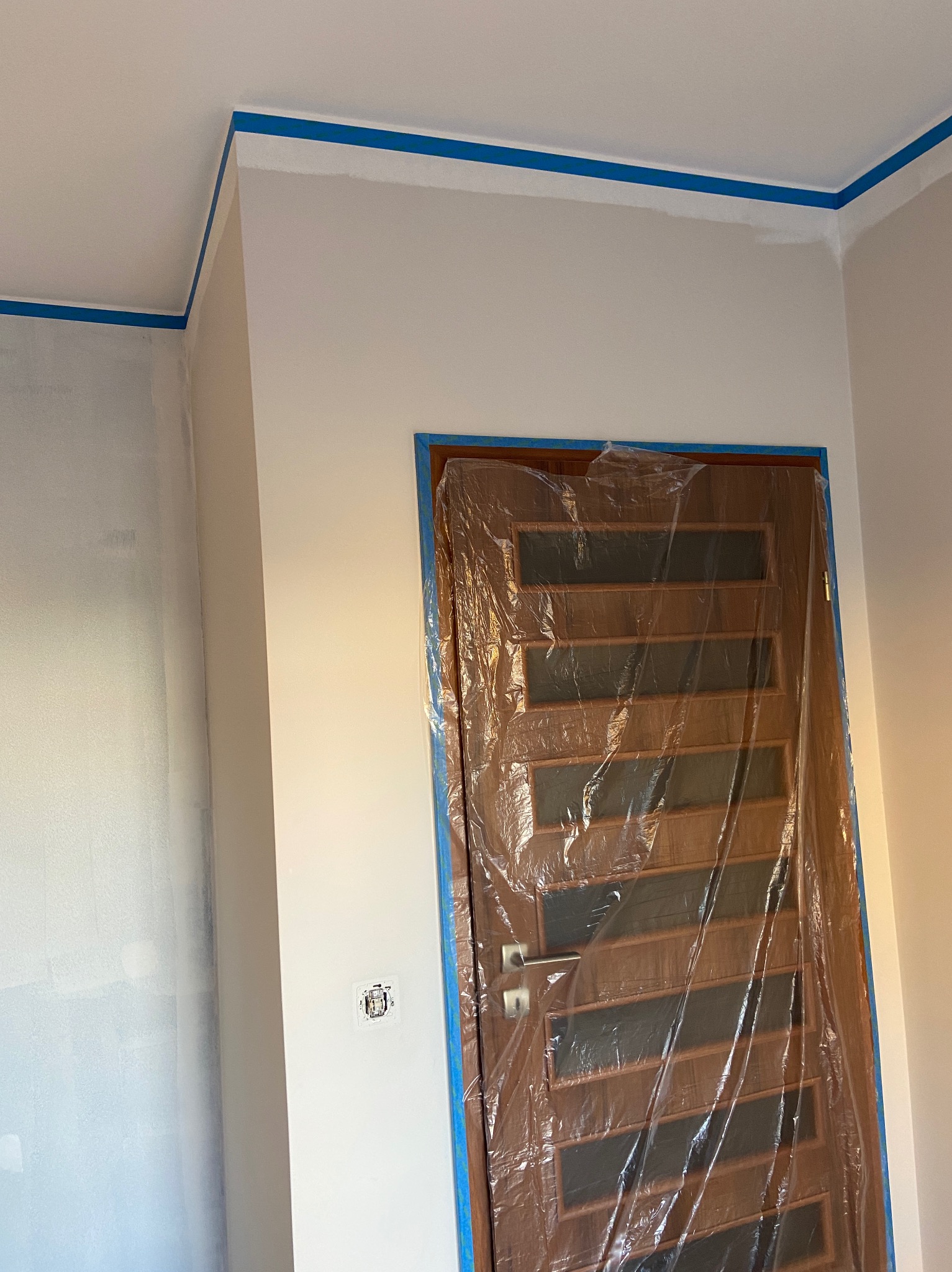 Przygotowanie do malowania: ściany zabezpieczone taśmą malarską przy suficie i narożnikach, drewniane drzwi oklejone folią ochronną, widoczny włącznik światła.