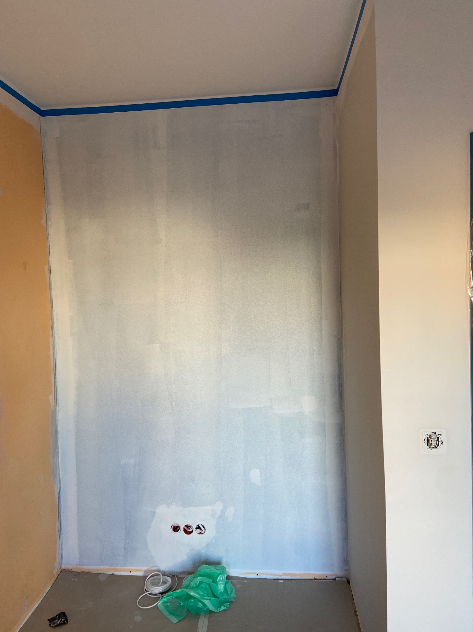 Przygotowana do malowania ściana z niebieską taśmą malarską przy suficie, widoczne puszki elektryczne i kabel na podłodze w folii.