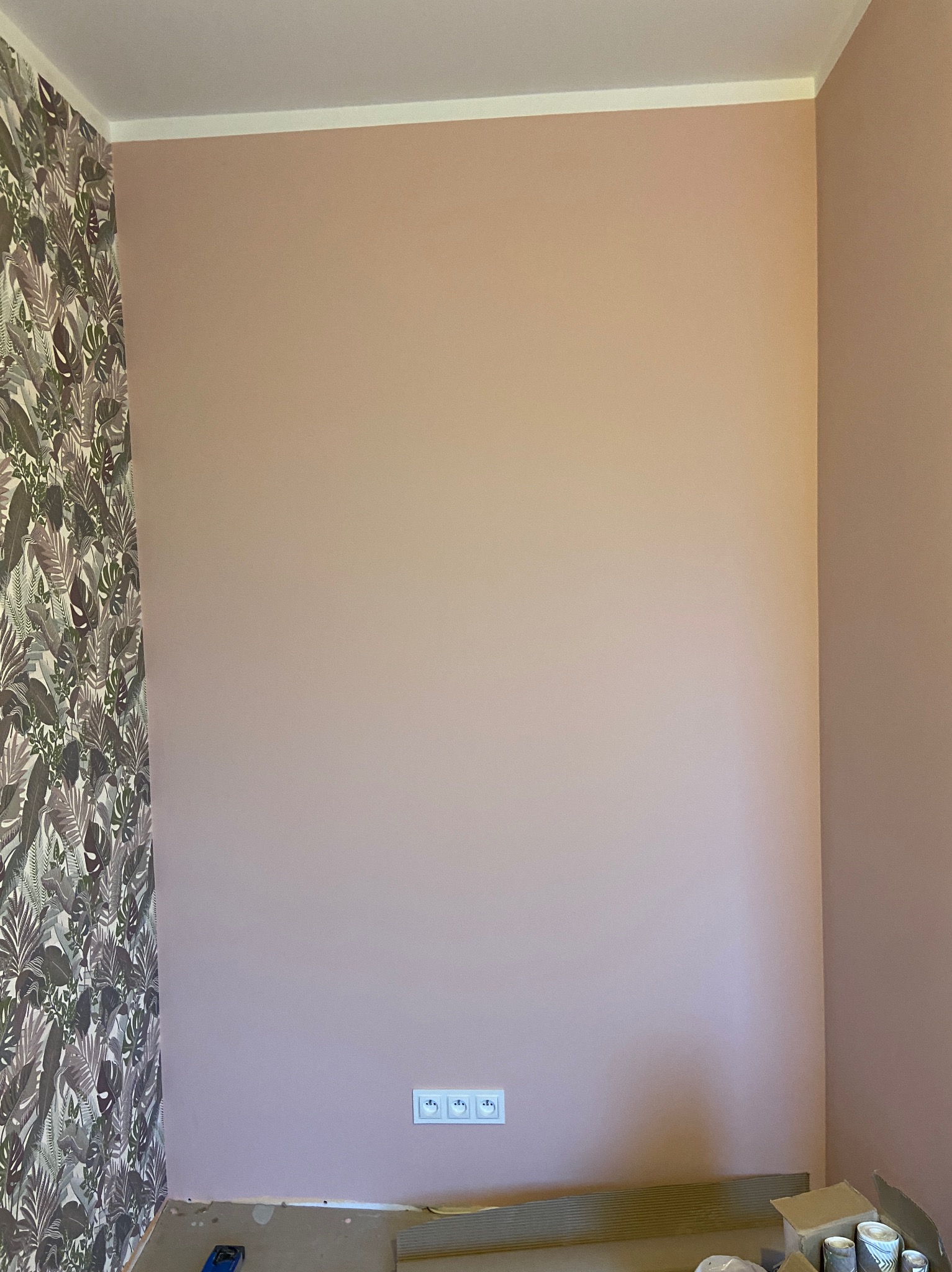 Ściana pomalowana na pudrowy róż z widocznymi gniazdkami elektrycznymi, fragmentem ściany z tapetą w liście oraz materiałami i narzędziami budowlanymi na podłodze, sugerującymi prace wykończeniowe.