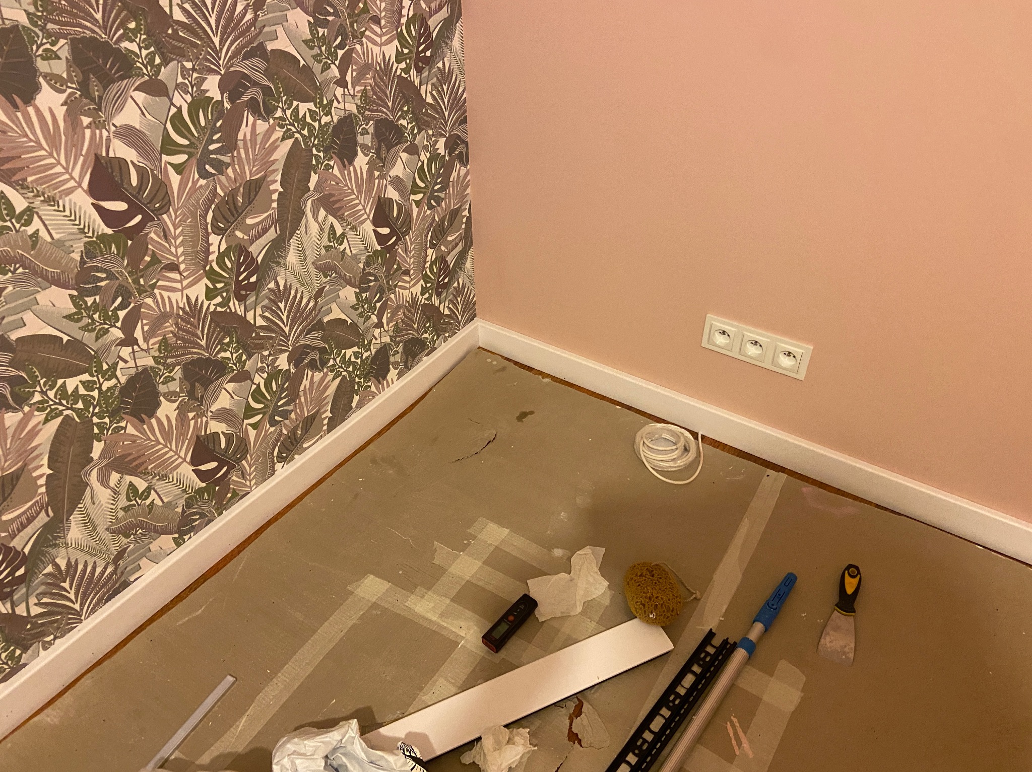 Pokój w trakcie odświeżania: ściana z tapetą w liście i różowa ściana z gniazdkami, podłoga przygotowana do położenia paneli, widoczne narzędzia, listwy przypodłogowe i zwój kabla.