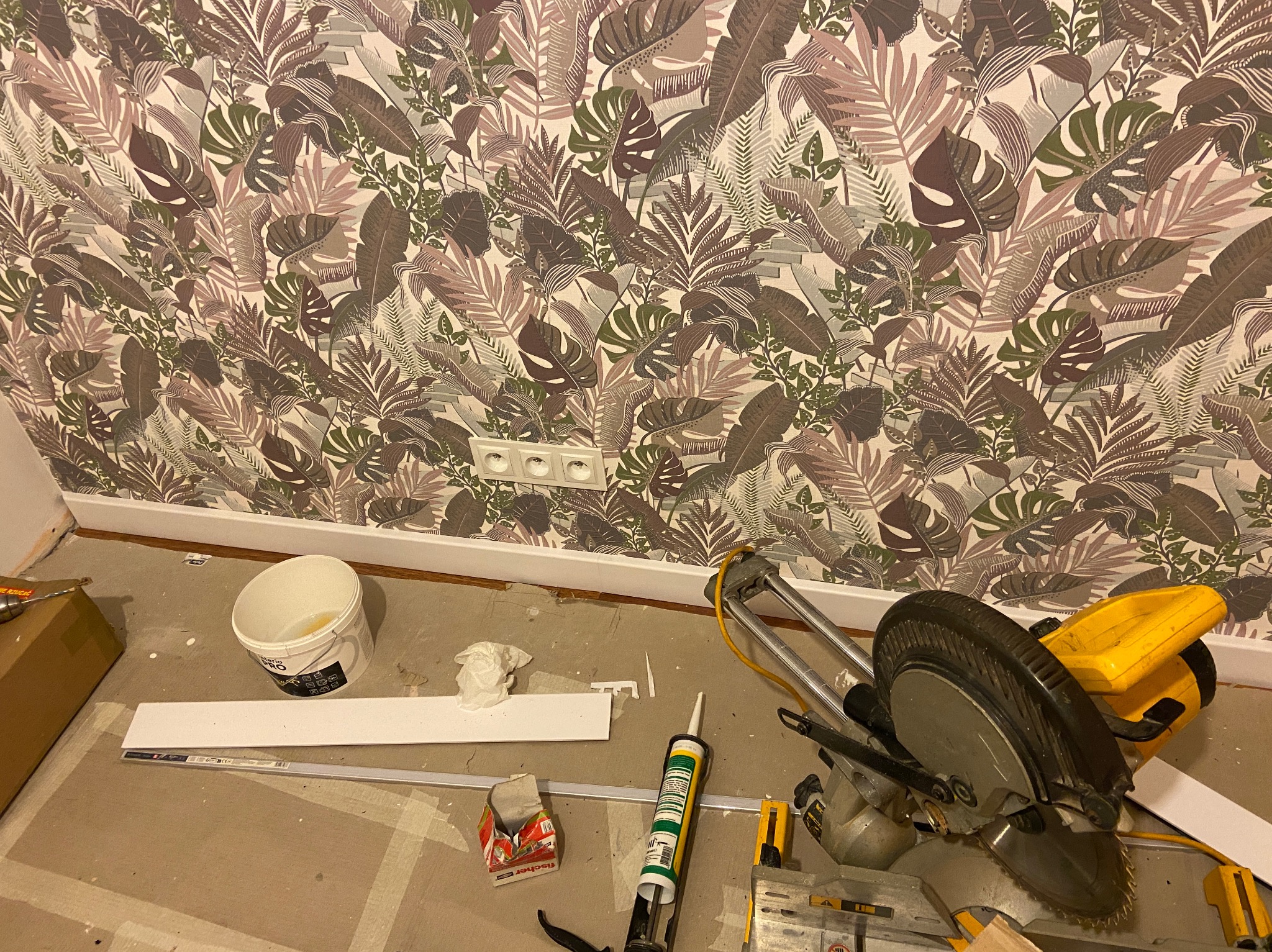 Pokój z tropikalną tapetą w odcieniach brązu i zieleni w trakcie wykańczania, widoczna biała listwa przypodłogowa, puszka farby, tuba z klejem i piła do cięcia.