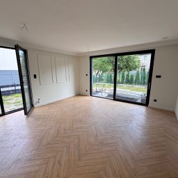Silesia Vision Mateusz Drzazgowski - Jasne wnętrze z parkietem ułożonym w jodełkę, duże okna przesuwne z widokiem na zieleń, białe ściany z dekoracyjnymi panelami. Częściowo widoczne rusztowanie za oknem.