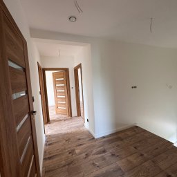 Silesia Vision Mateusz Drzazgowski - Jasne wnętrze po remoncie: drewniane panele podłogowe, świeżo pomalowane białe ściany, zamontowane drewniane drzwi z przeszkleniami. Widok z perspektywy korytarza.