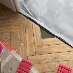 Silesia Vision Mateusz Drzazgowski - Układanie parkietu w jodełkę, widoczne nakolanniki, taśma malarska i folia ochronna. Fragment podłogi przed i po ułożeniu paneli. Praca glazurnika w toku.