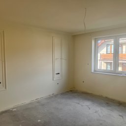 Silesia Vision Mateusz Drzazgowski - Pokój po gładzi, z dekoracyjnymi ramami na ścianach i widocznymi otworami na gniazdka. Surowa podłoga betonowa, białe okno. Oświetlenie dzienne.