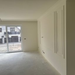 Silesia Vision Mateusz Drzazgowski - Jasne wnętrze po remoncie: świeżo pomalowane ściany z dekoracyjnymi listwami, duże okno balkonowe z widokiem na dom, surowa betonowa podłoga, przygotowane do dalszych prac wykończeniowych.