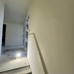 Silesia Vision Mateusz Drzazgowski - Wnętrze w trakcie prac wykończeniowych: gładkie, jasne ściany, surowy beton na podłodze, widoczne kable i lampa budowlana. Otwór drzwiowy z widoczną konstrukcją.