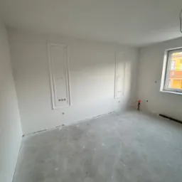 Wnętrze pokoju po gładziowaniu ścian z dekoracyjnymi ramkami wokół gniazdek elektrycznych. Szare podłoże pod przyszłą podłogę, okno z widokiem na budynek.