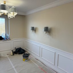 Silesia Vision Mateusz Drzazgowski - Pokój w trakcie malowania z zabezpieczonymi lampami i listwami, widoczne narzędzia malarskie i wiadro z farbą. Podłoga zabezpieczona folią malarską.