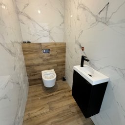Silesia Vision Mateusz Drzazgowski - Nowoczesna łazienka z marmurowymi płytkami ściennymi, drewnopodobną podłogą i wiszącą toaletą. Czarna szafka z białą umywalką i czarną baterią.