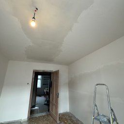 Silesia Vision Mateusz Drzazgowski - Pokój w trakcie malowania: ściany i sufit częściowo pokryte farbą, drabina oparta o ścianę, widoczne ślady farby na podłodze, otwarte drzwi do kolejnego pomieszczenia.