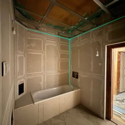 Silesia Vision Mateusz Drzazgowski - Łazienka w trakcie remontu: zabudowa wanny płytami, widoczne profile sufitowe i zielona linia lasera wyznaczająca poziom. Otwór drzwiowy z widokiem na materiały budowlane.