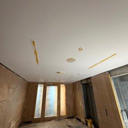 Silesia Vision Mateusz Drzazgowski - Wnętrze w trakcie prac wykończeniowych: zabezpieczone folią ściany, sufit po gładziowaniu z widocznymi miejscami po instalacjach i taśmami malarskimi.