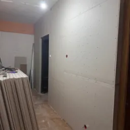 Montaż płyt gipsowo-kartonowych na ścianie w korytarzu, widoczne płyty czekające na montaż, instalacja elektryczna w ścianie. W tle widoczne drzwi i narzędzia.