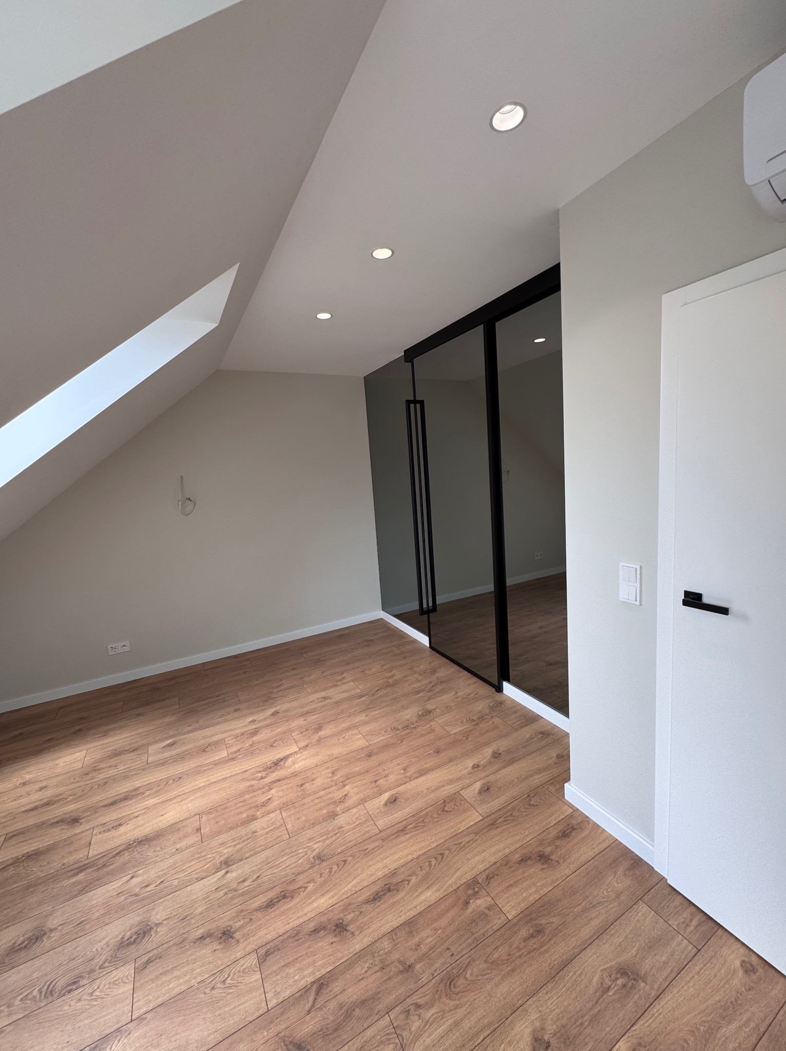 Pokój na poddaszu z brązowymi panelami, białymi ścianami i drzwiami oraz szafą z czarnymi ramami. Widoczne skosy sufitu i oświetlenie punktowe.