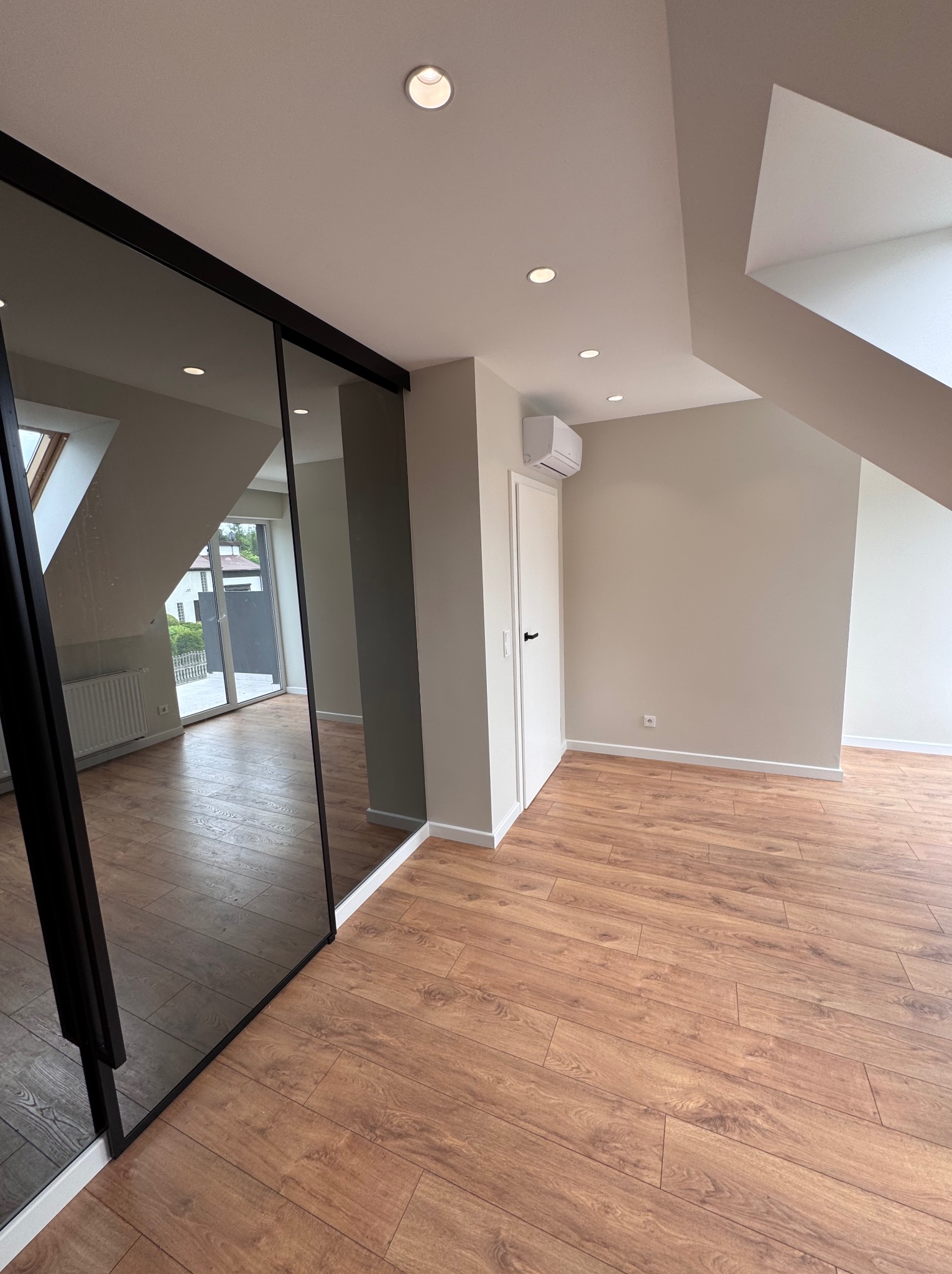 Jasne poddasze z drewnianą podłogą i przesuwną szafą z lustrzanymi drzwiami. Widoczne oświetlenie sufitowe i klimatyzator. Minimalistyczny design.