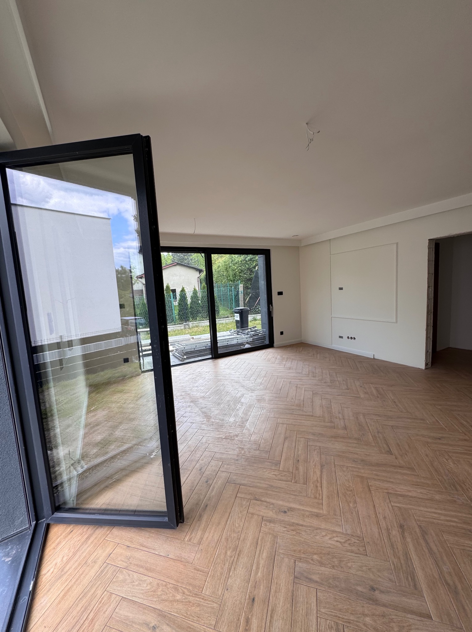 Jasne wnętrze z parkietem ułożonym w jodełkę, otwartym oknem tarasowym i minimalistycznymi dekoracjami ściennymi. Widoczne przygotowania do dalszych prac.