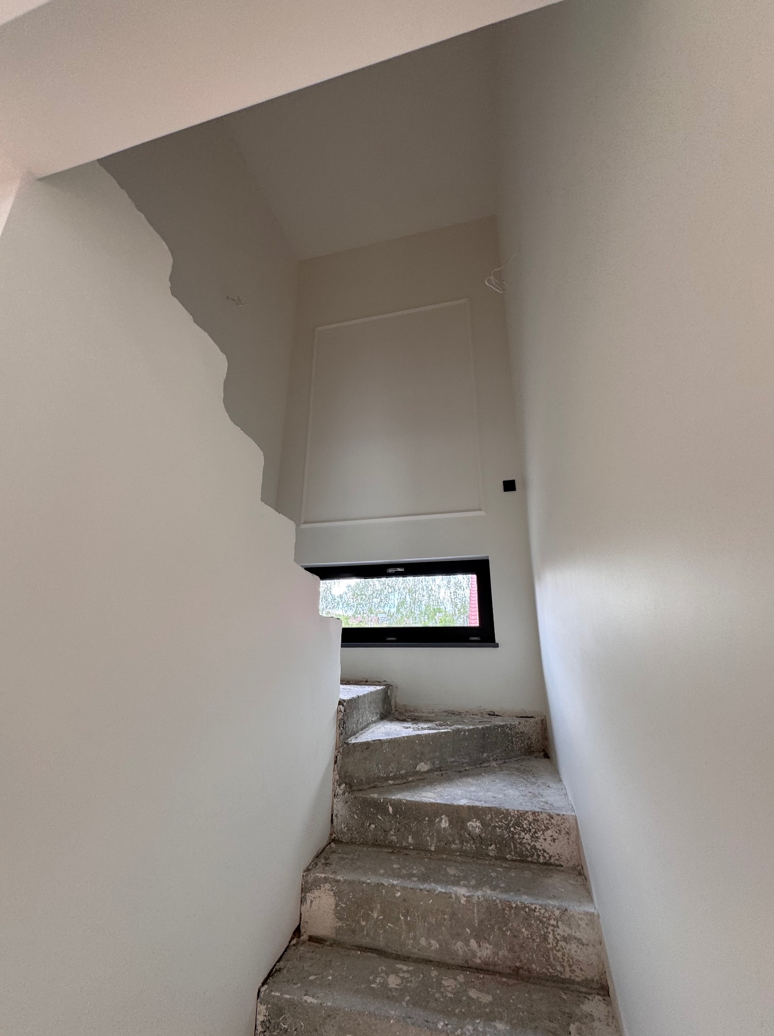 Wnętrze w trakcie remontu: surowe schody betonowe prowadzą w górę, ściany częściowo pokryte gładzią, małe okno w czarnej ramie. Widoczne niedoskonałości i etap prac wykończeniowych.