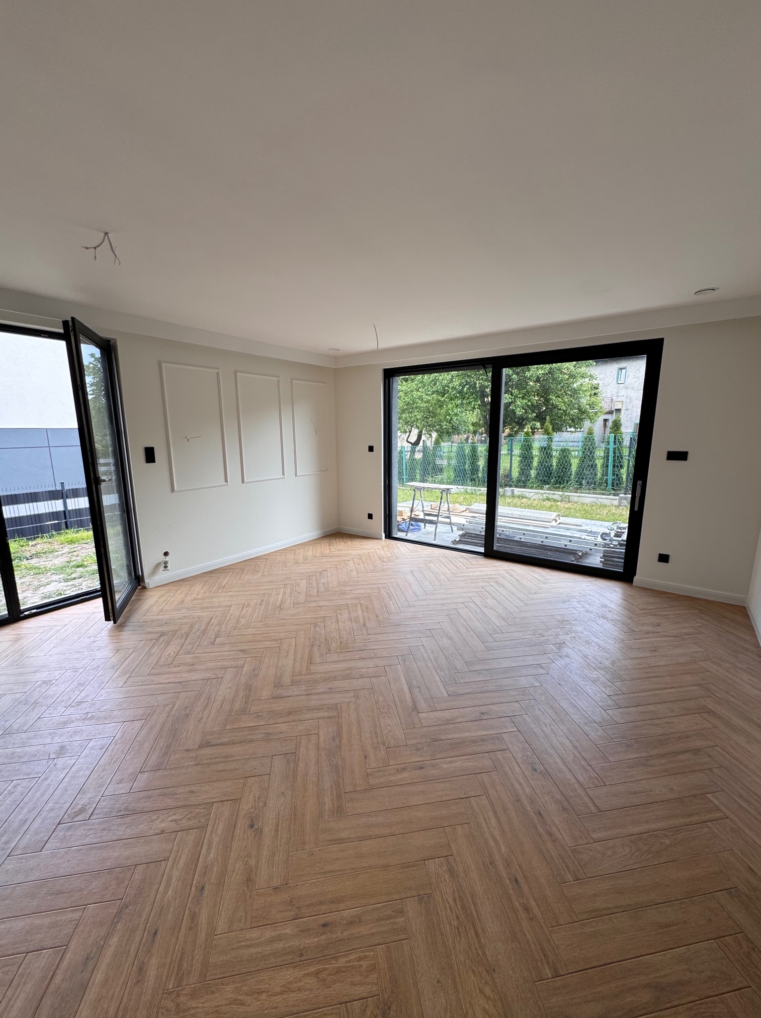 Jasne wnętrze z parkietem ułożonym w jodełkę, duże okna przesuwne z widokiem na zieleń, białe ściany z dekoracyjnymi panelami. Częściowo widoczne rusztowanie za oknem.