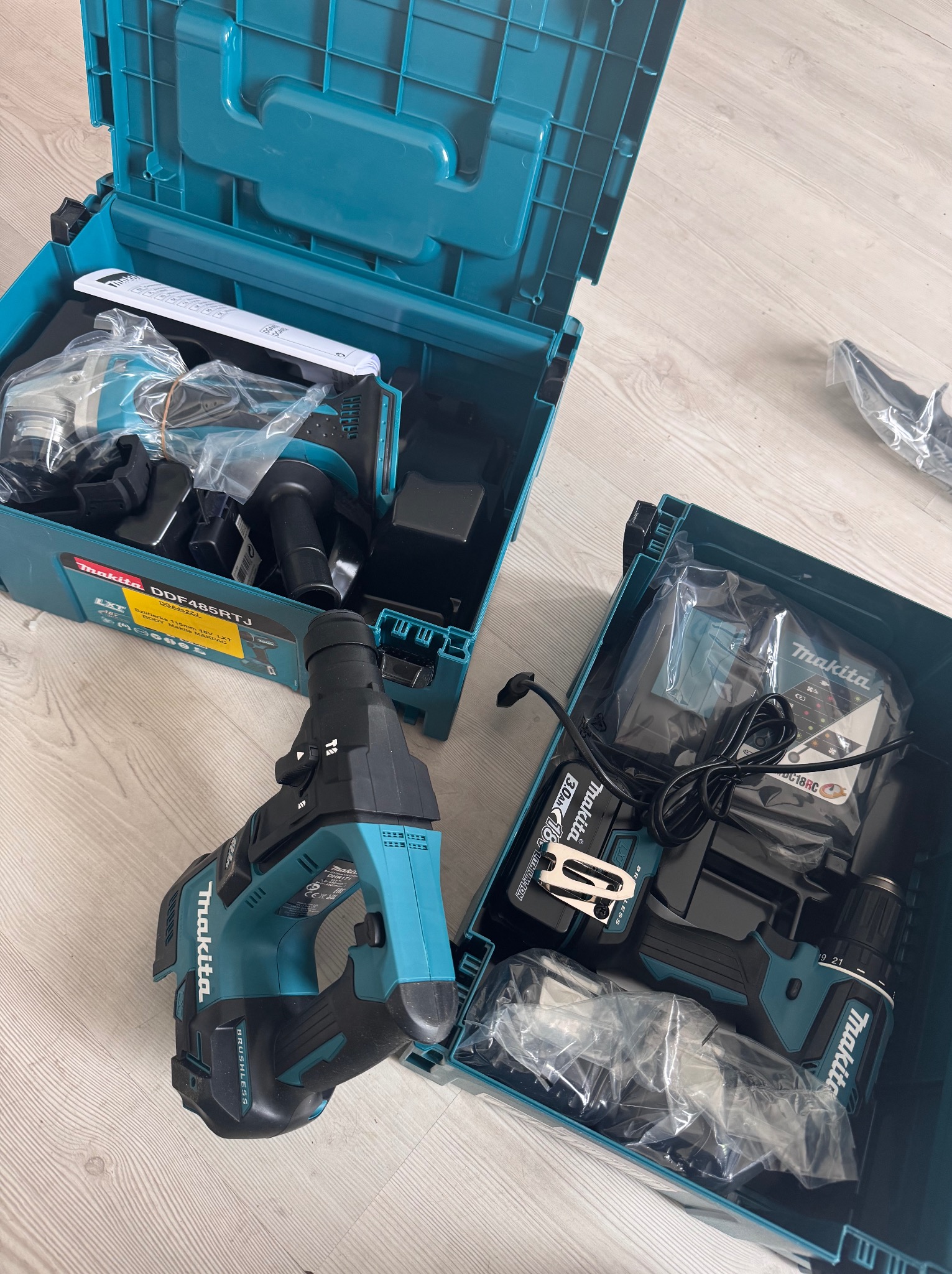 Zestaw narzędzi akumulatorowych Makita w turkusowych skrzynkach. Wiertarko-wkrętarka i młot udarowy, ładowarka oraz akumulatory na jasnej podłodze.