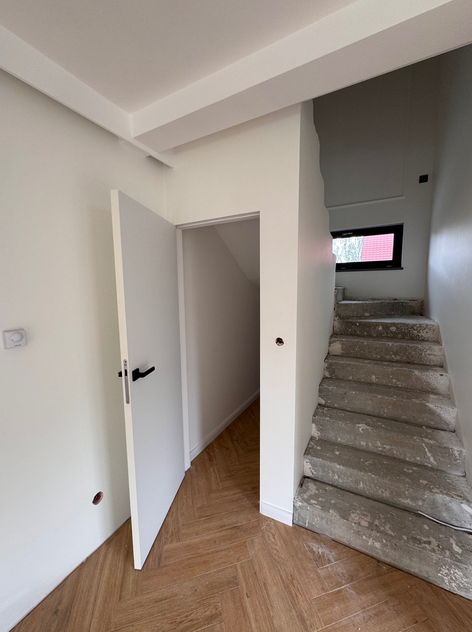 Wnętrze w trakcie prac: białe ściany, otwarte drzwi z czarną klamką, surowe betonowe schody i parkiet w jodełkę. Widoczne niedokończone prace wykończeniowe.