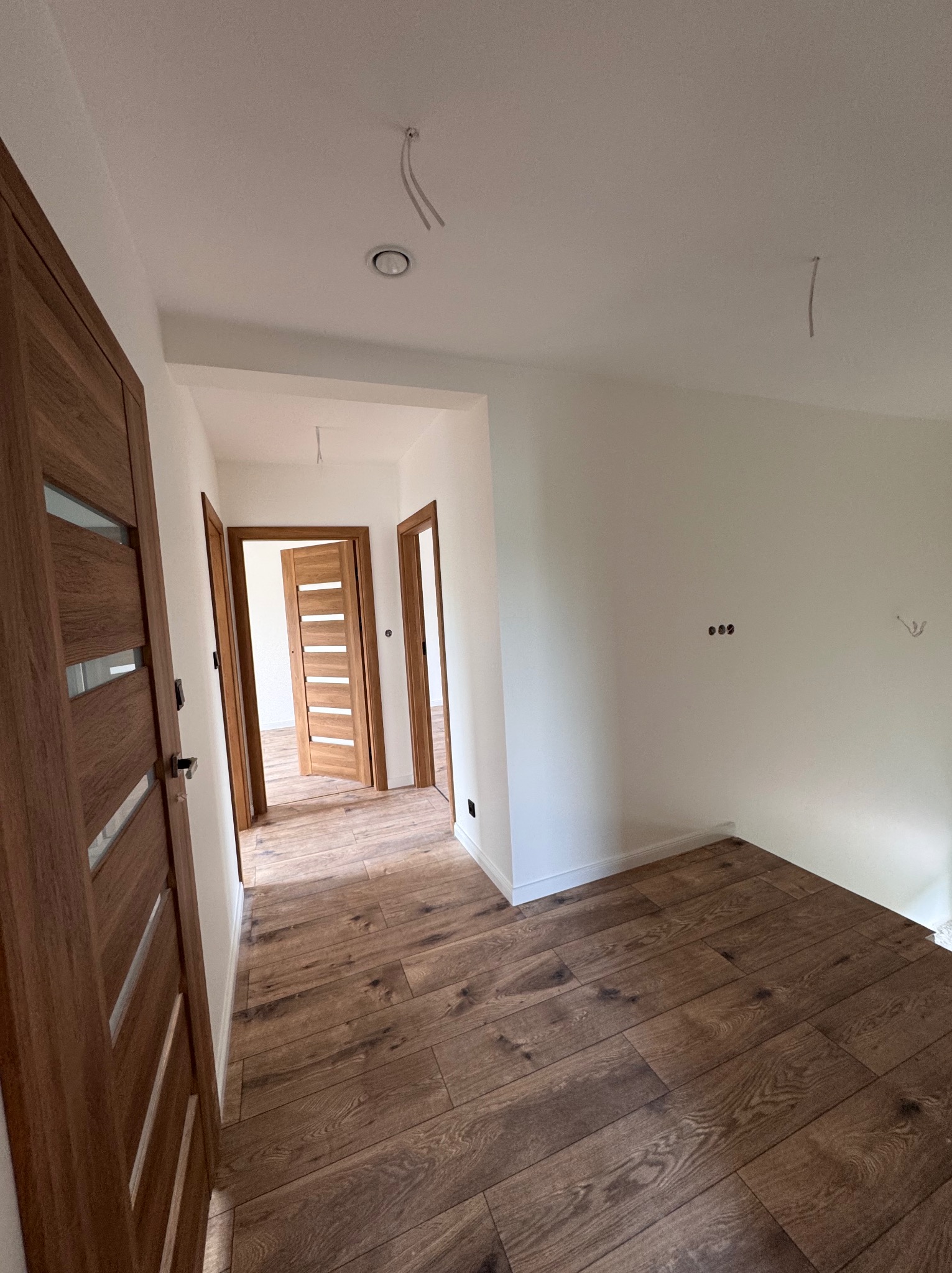 Jasne wnętrze po remoncie: drewniane panele podłogowe, świeżo pomalowane białe ściany, zamontowane drewniane drzwi z przeszkleniami. Widok z perspektywy korytarza.
