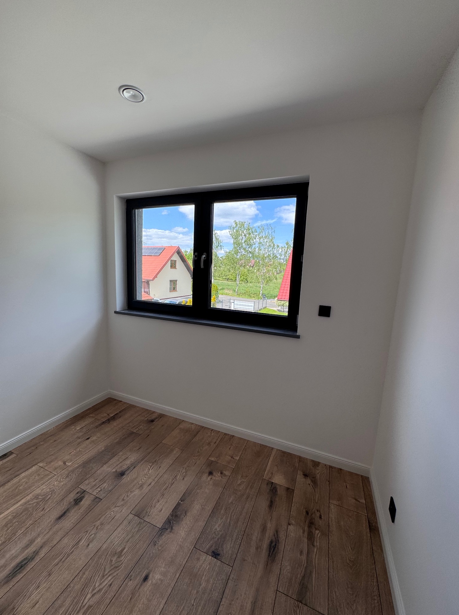 Jasne wnętrze po remoncie: drewniana podłoga, białe ściany, czarna rama okna z widokiem na dom i zieleń. Minimalistyczny design i nowoczesne wykończenie.