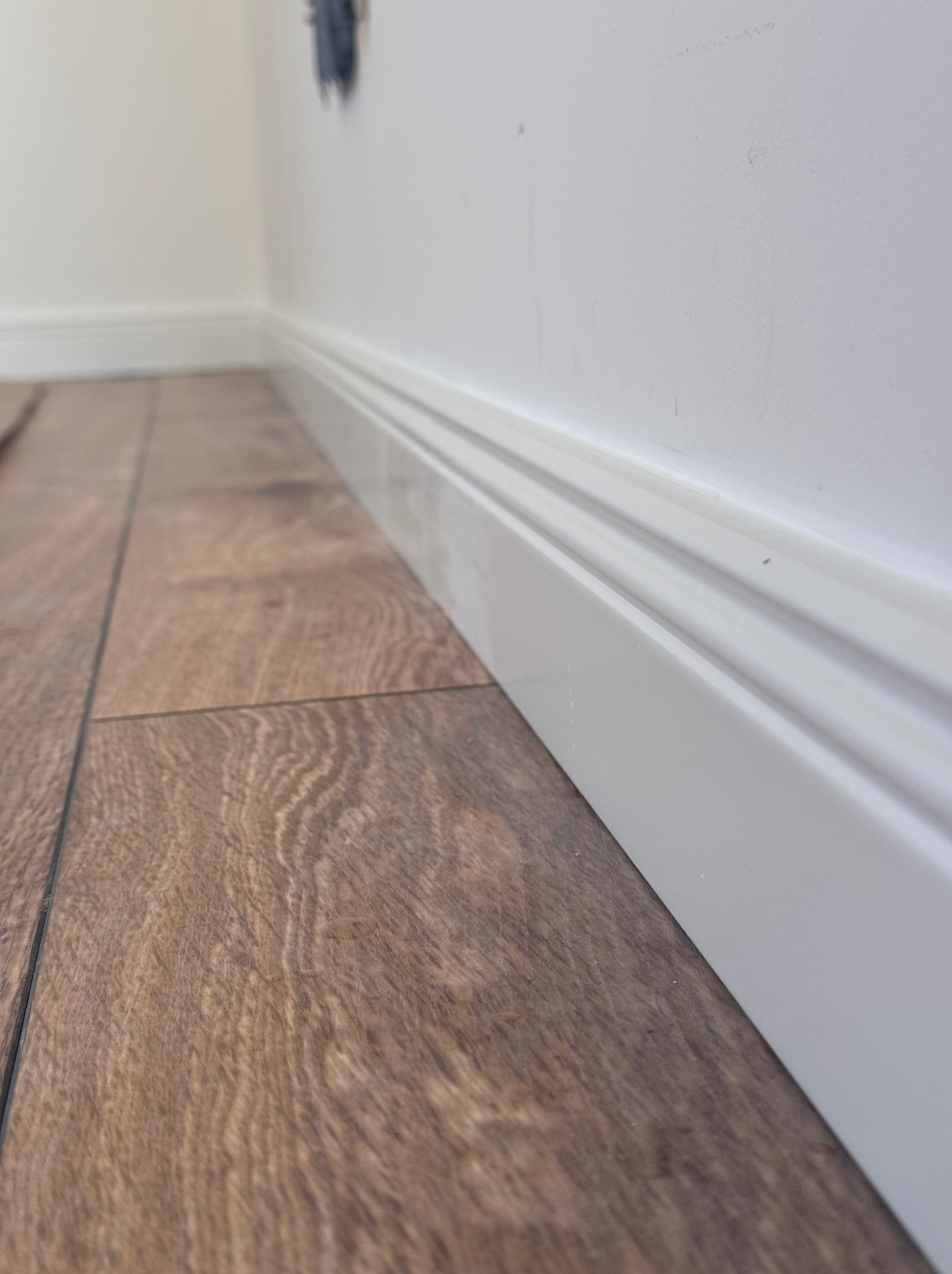 Ułożone panele podłogowe w odcieniach brązu, z białą listwą przypodłogową i fragmentem białej ściany z gniazdkiem. Niski kąt ujęcia podkreśla fakturę paneli.