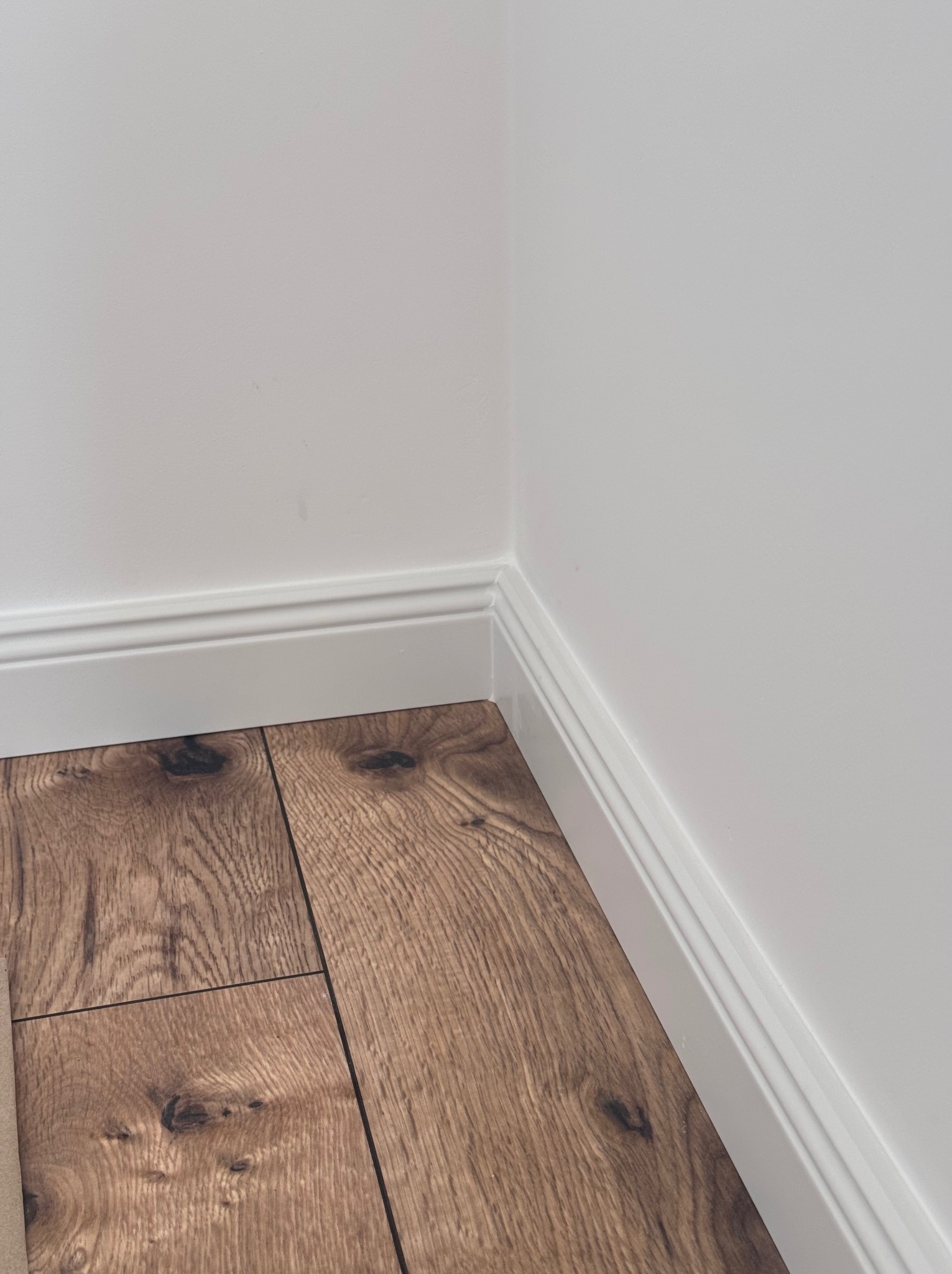 Fragment wykończonego wnętrza: brązowe panele podłogowe imitujące drewno, białe ściany z listwami przypodłogowymi, narożnik pokoju. Ujęcie z dołu.