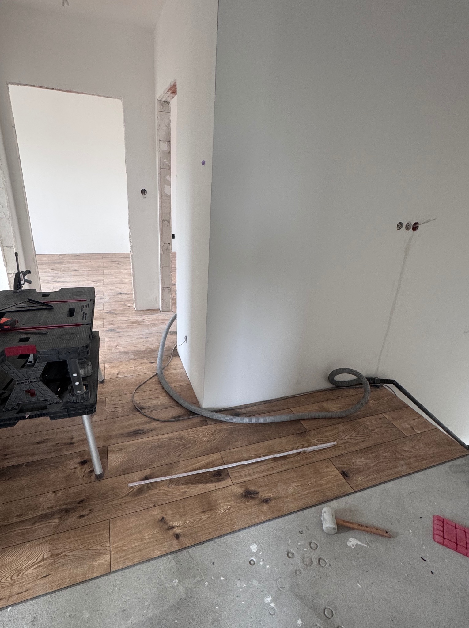 Układanie paneli podłogowych z wzorem drewna w jasnym wnętrzu. Widoczny stół roboczy, narzędzia i fragment ściany z instalacją elektryczną. Podłoga częściowo wykończona.