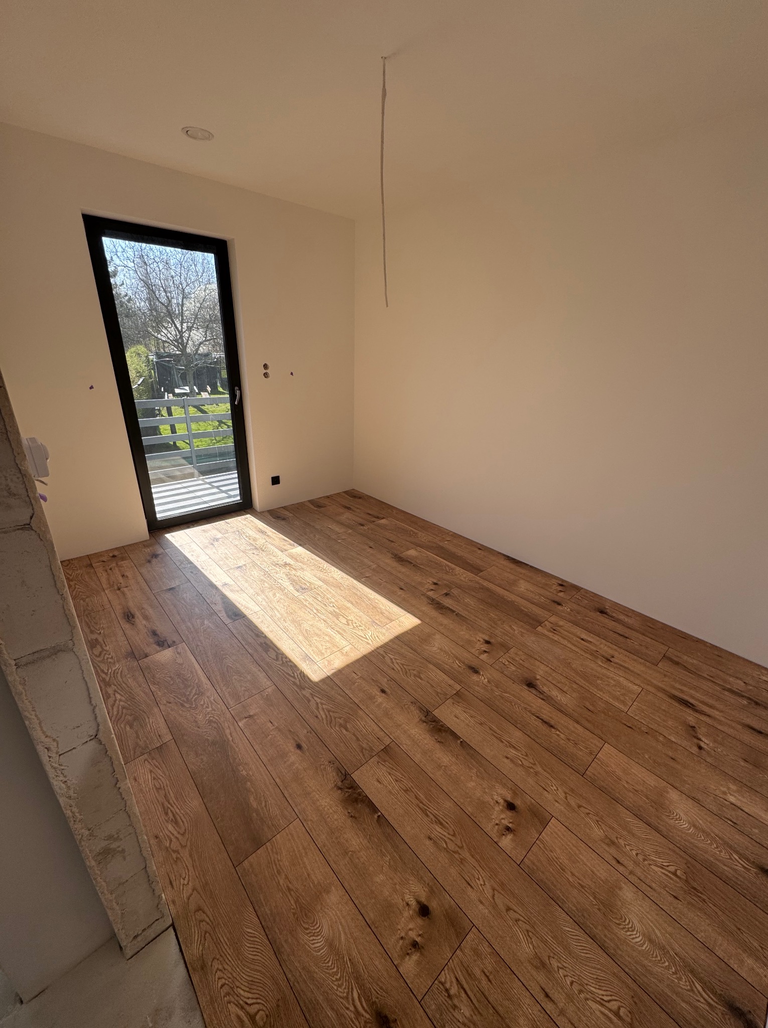 Jasny pokój z ułożonymi panelami podłogowymi w odcieniach brązu, widok na balkon przez czarne ramy okienne. Świeże panele podłogowe.