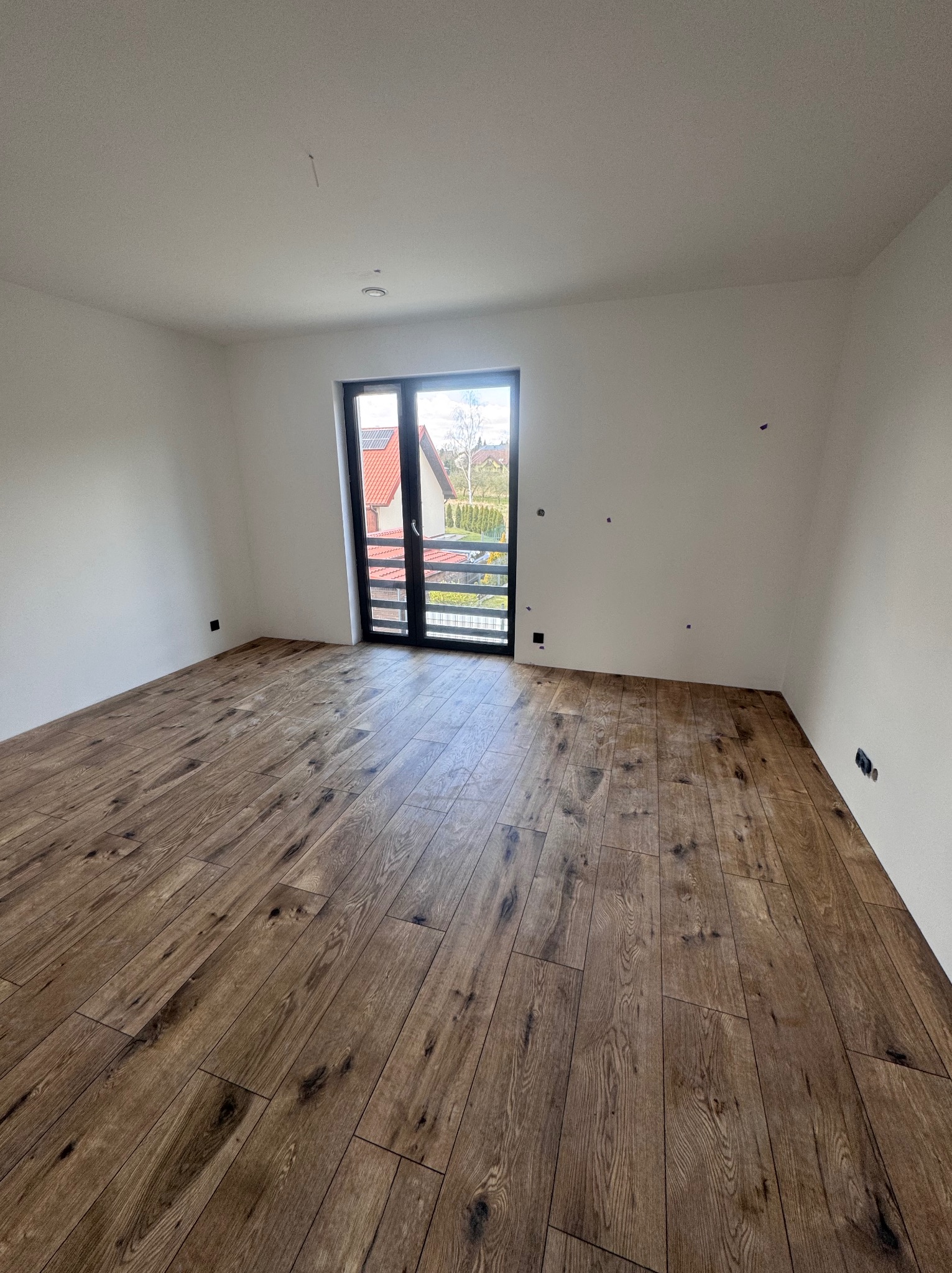 Pokój po remoncie z panelami podłogowymi w odcieniach brązu i białymi ścianami. Widoczne ślady po taśmie malarskiej i okno balkonowe z czarną ramą.