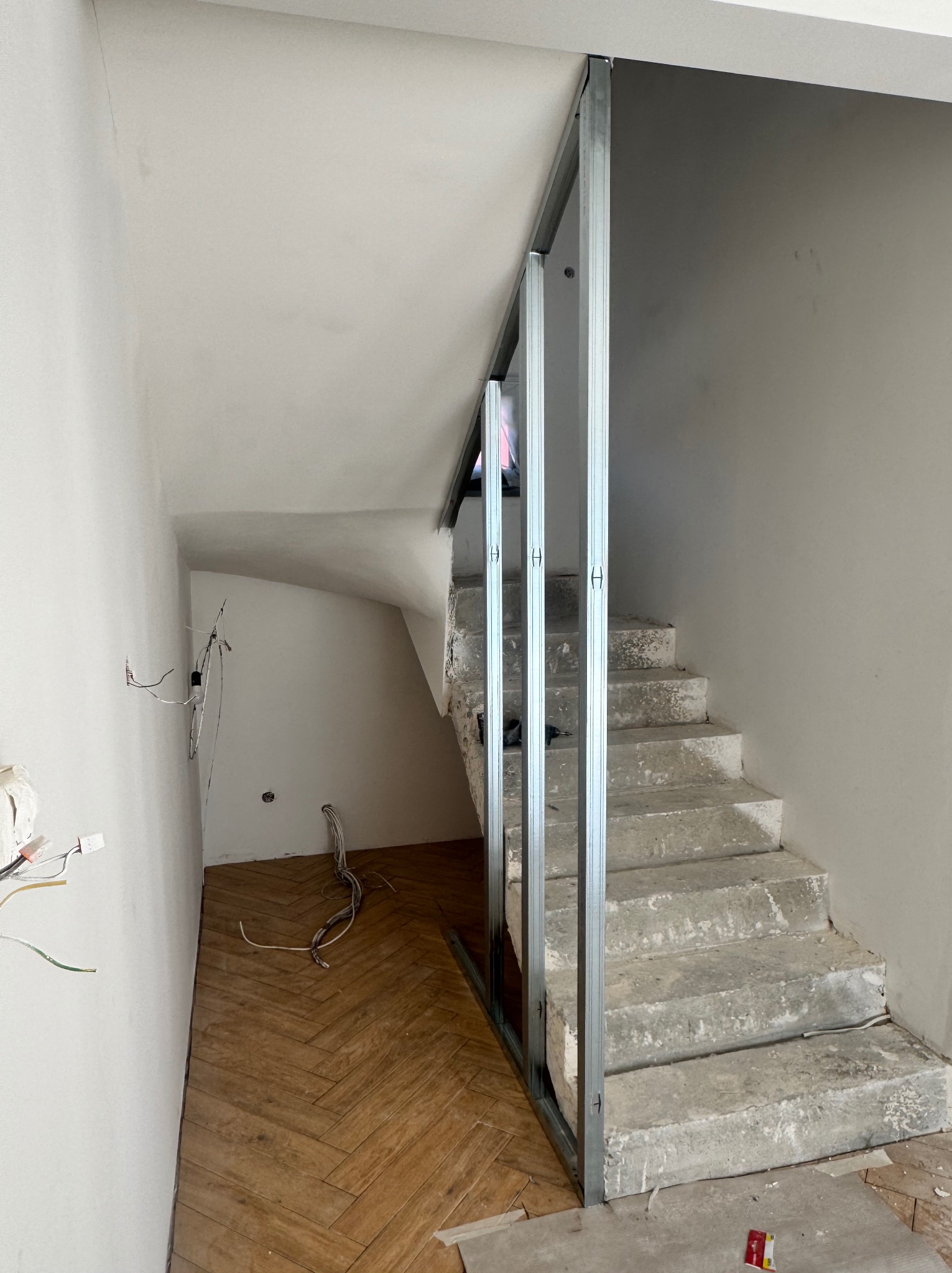 Wnętrze w trakcie prac: metalowa konstrukcja ścianki działowej przy schodach, widoczne przewody elektryczne i parkiet ułożony w jodełkę na podłodze.