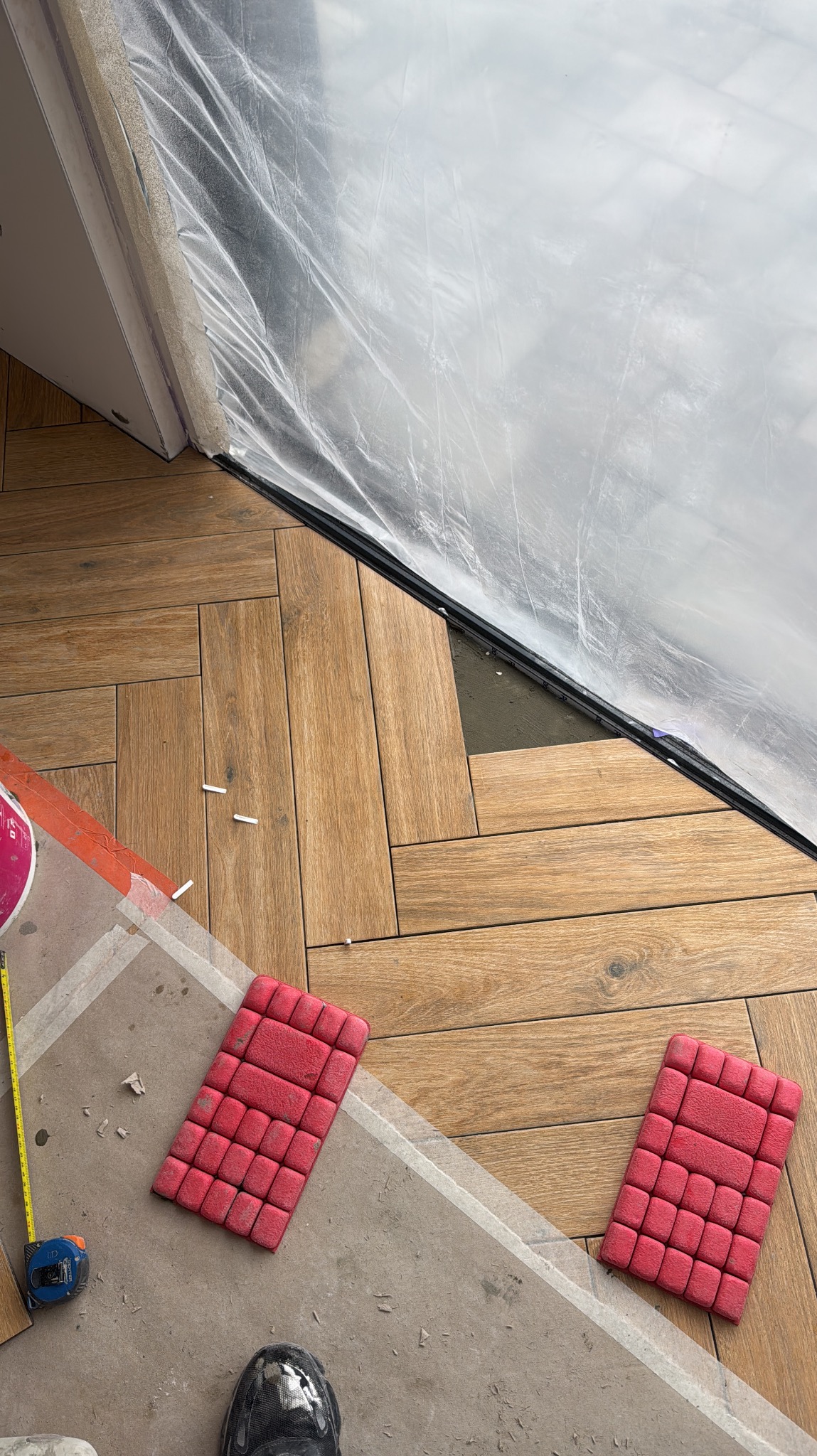 Układanie parkietu w jodełkę, widoczne nakolanniki, taśma malarska i folia ochronna. Fragment podłogi przed i po ułożeniu paneli. Praca glazurnika w toku.