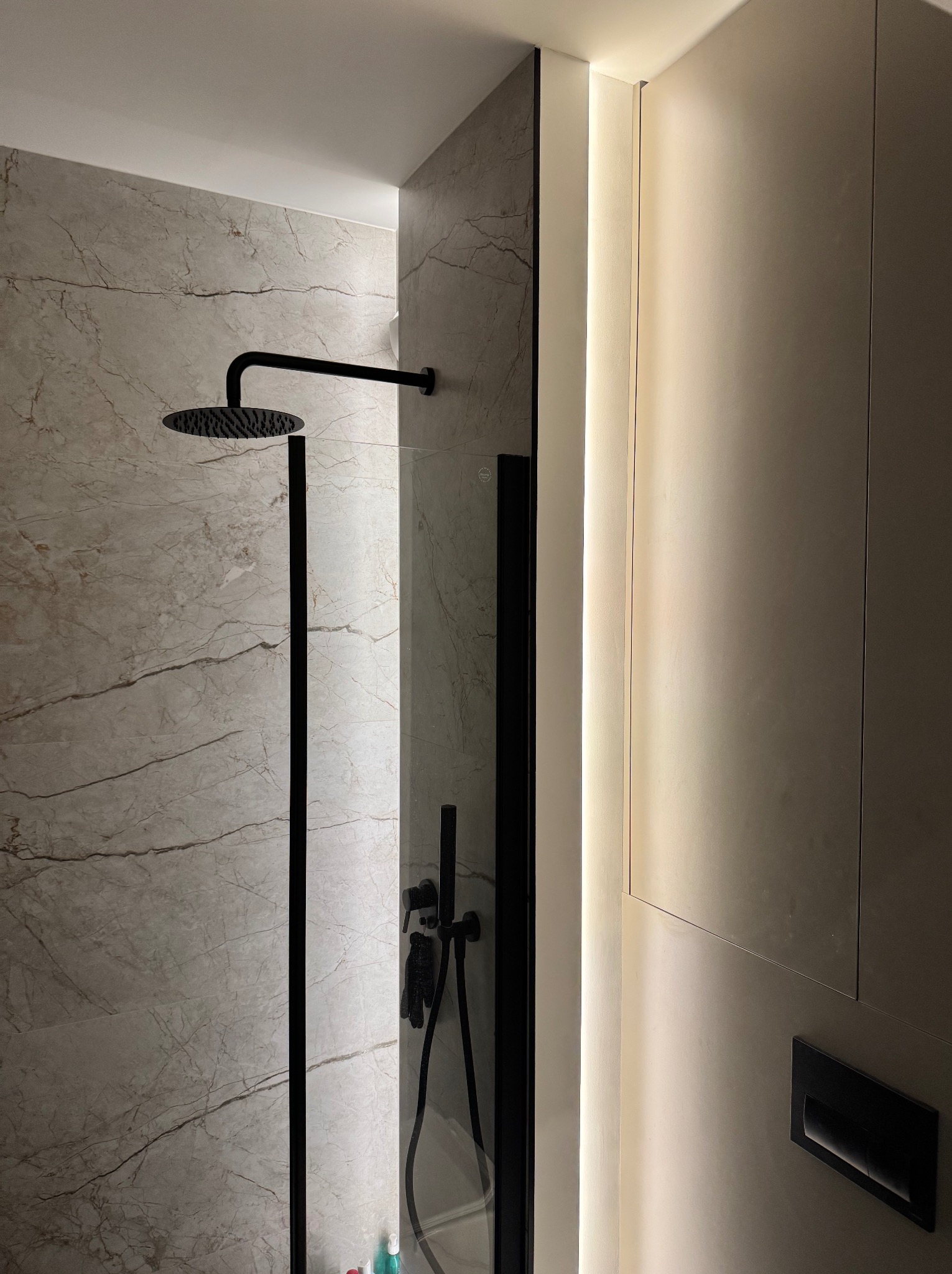 Nowoczesna kabina prysznicowa z deszczownicą i szklaną ścianką, podświetlona listwa LED. Marmurowe płytki i czarna armatura.
