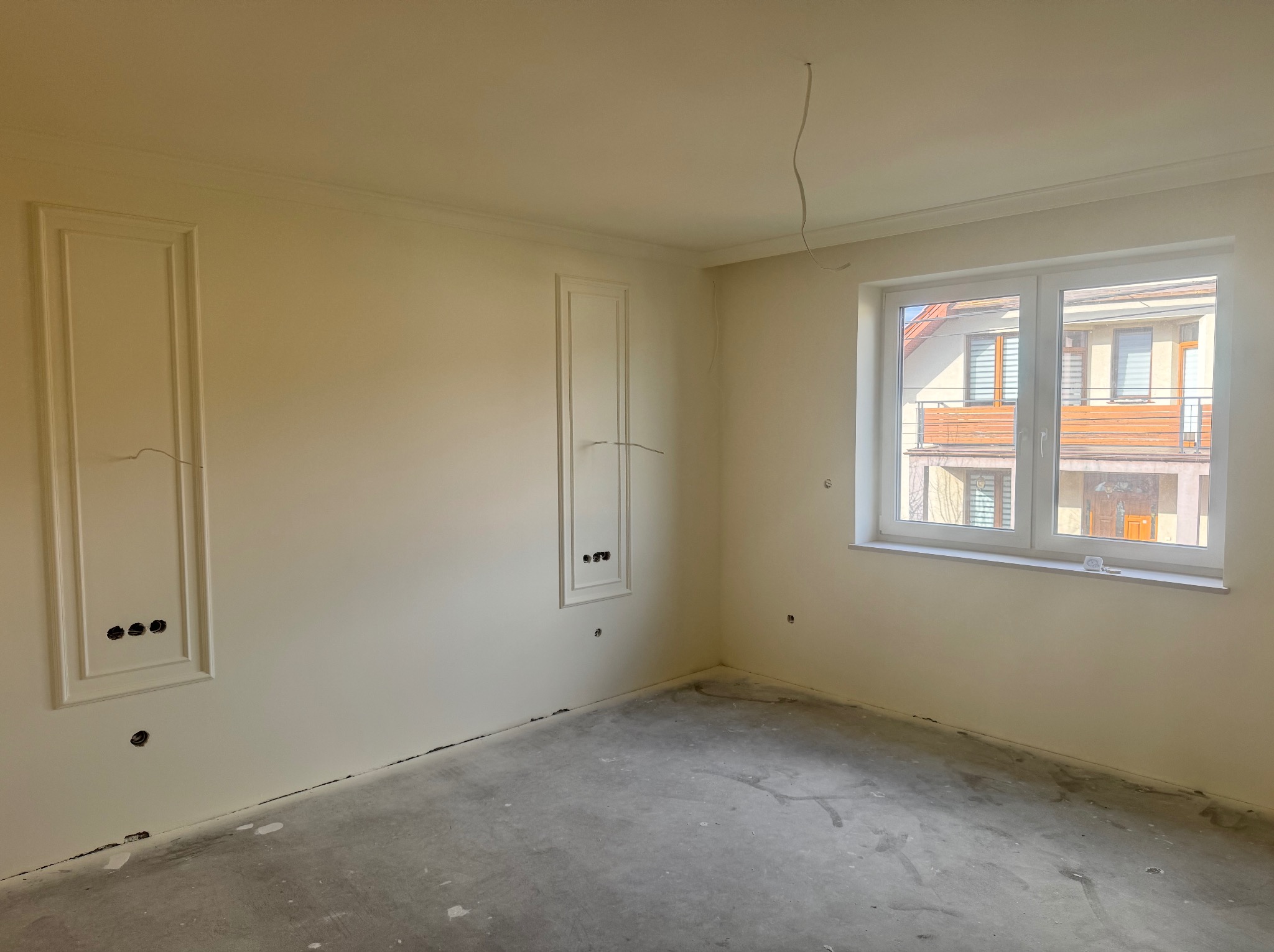 Pokój po gładzi, z dekoracyjnymi ramami na ścianach i widocznymi otworami na gniazdka. Surowa podłoga betonowa, białe okno. Oświetlenie dzienne.