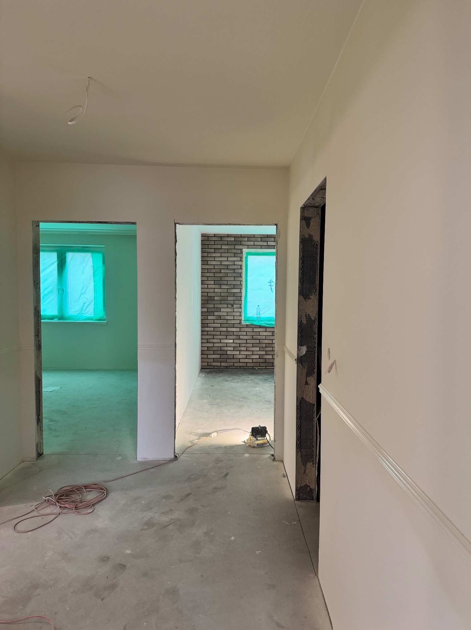 Wnętrze w trakcie prac wykończeniowych: białe ściany z dekoracyjną listwą, surowy beton na podłodze, widoczne przygotowania do montażu drzwi, okna zabezpieczone folią.