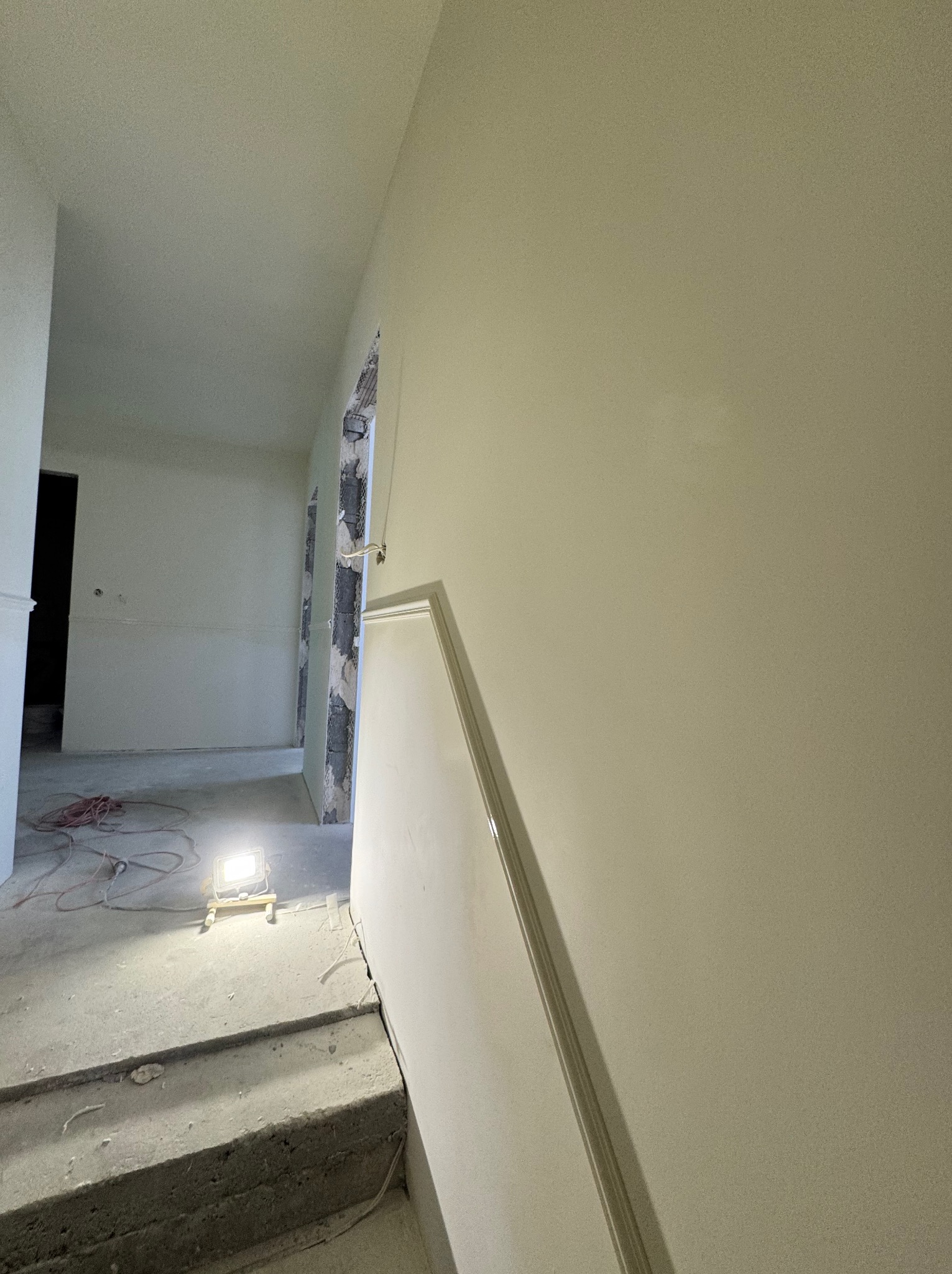 Wnętrze w trakcie prac wykończeniowych: gładkie, jasne ściany, surowy beton na podłodze, widoczne kable i lampa budowlana. Otwór drzwiowy z widoczną konstrukcją.