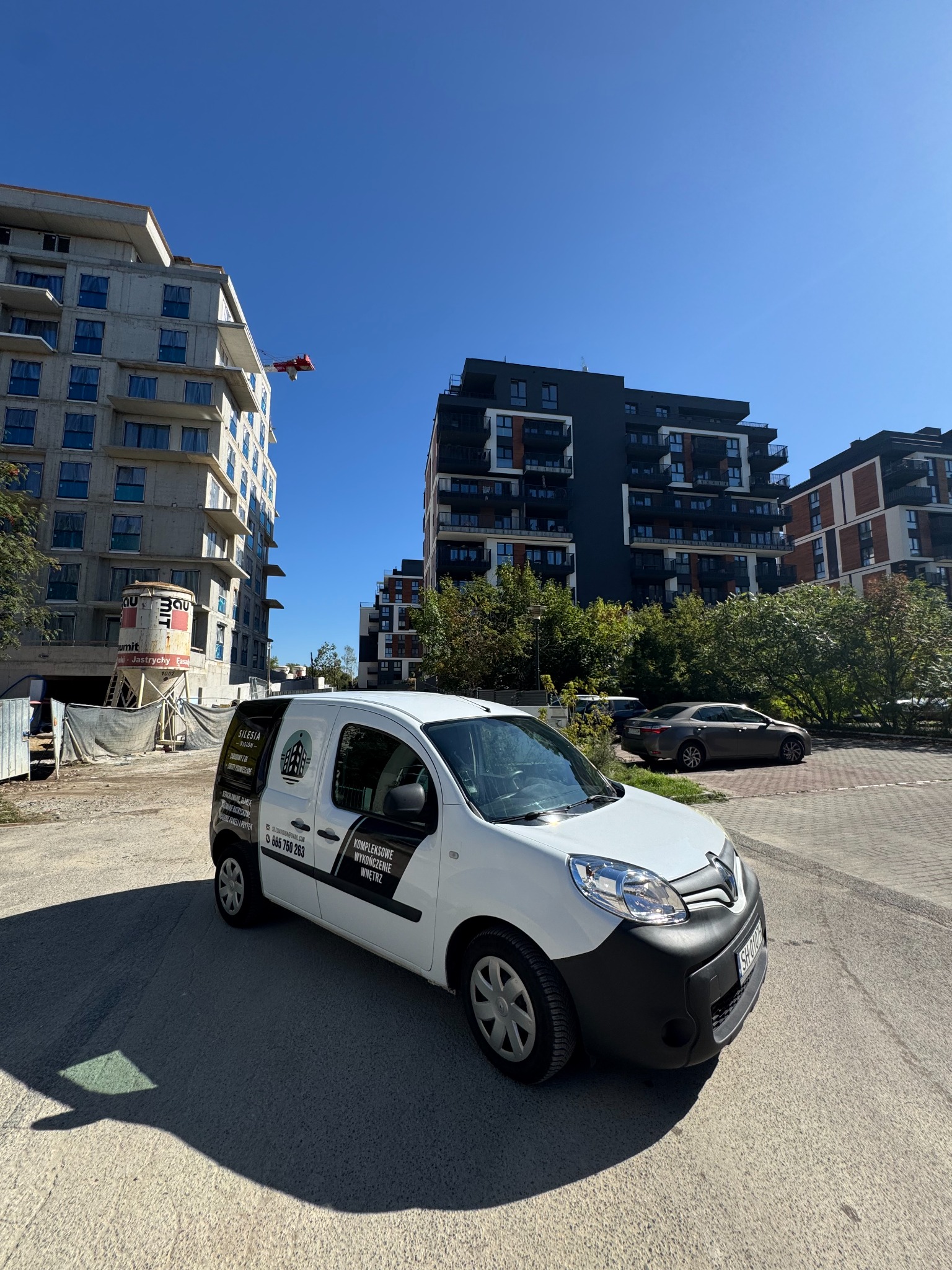 Biały samochód dostawczy z logo firmy remontowej na tle budynków w trakcie budowy i gotowych apartamentowców w słoneczny dzień.