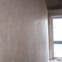 Świeżo nałożona warstwa jasnego tynku na ścianie w surowym wnętrzu, widoczna faktura i nieregularności, okno w tle z resztkami gruzu na parapecie.