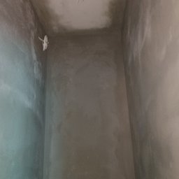 Przemysław Kuras PRZEM-BUD - Wnętrze pomieszczenia w trakcie prac wykończeniowych; ściany pokryte świeżym, szarym tynkiem. Widoczne przewody elektryczne wystające ze ściany i sufitu.