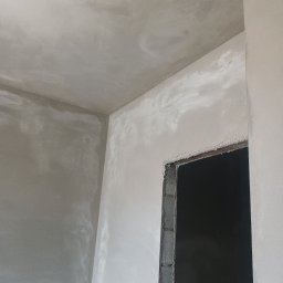 Przemysław Kuras PRZEM-BUD - Wnętrze w trakcie prac wykończeniowych, surowe ściany pokryte świeżym, jasnoszarym tynkiem, widoczne nierówności i zacieki, sufit również otynkowany, w tle otwór drzwiowy z surową ramą.