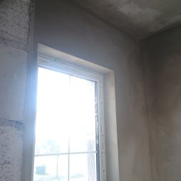 Przemysław Kuras PRZEM-BUD - Świeżo otynkowane ściany w narożniku pomieszczenia z oknem, widoczny fragment surowego muru przy ościeżnicy okiennej.