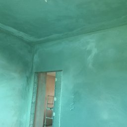 Przemysław Kuras PRZEM-BUD - Świeżo nałożona, wilgotna, zielonkawa warstwa tynku na ścianach i suficie, widoczna drabina w przejściu drzwiowym.