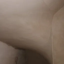 Świeżo położona, gładka, beżowa warstwa tynku na suficie i ścianach z zaokrąglonym narożnikiem, widoczne drobne niedoskonałości i ślady po narzędziach.