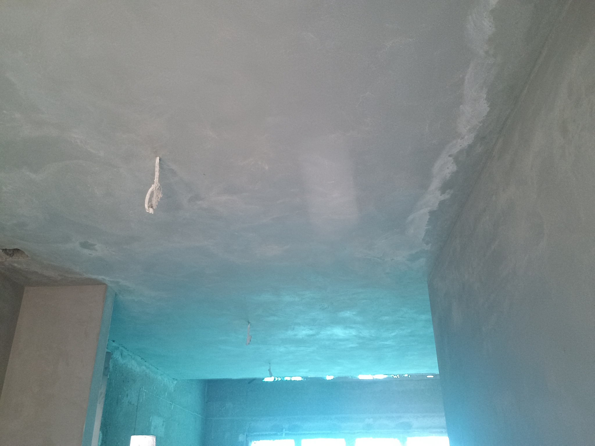 Świeżo nałożony, szary tynk na suficie w trakcie schnięcia, widoczne nierówności i fragmenty wystających kabli, w tle otwór okienny z intensywnym światłem dziennym.