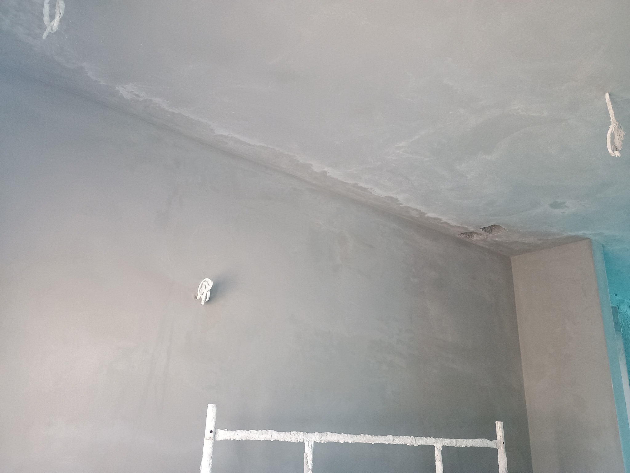 Świeżo nałożona, szara, gładka powierzchnia tynku na ścianach i suficie w narożnym pokoju, widoczne wyprowadzone kable elektryczne i fragment rusztowania.